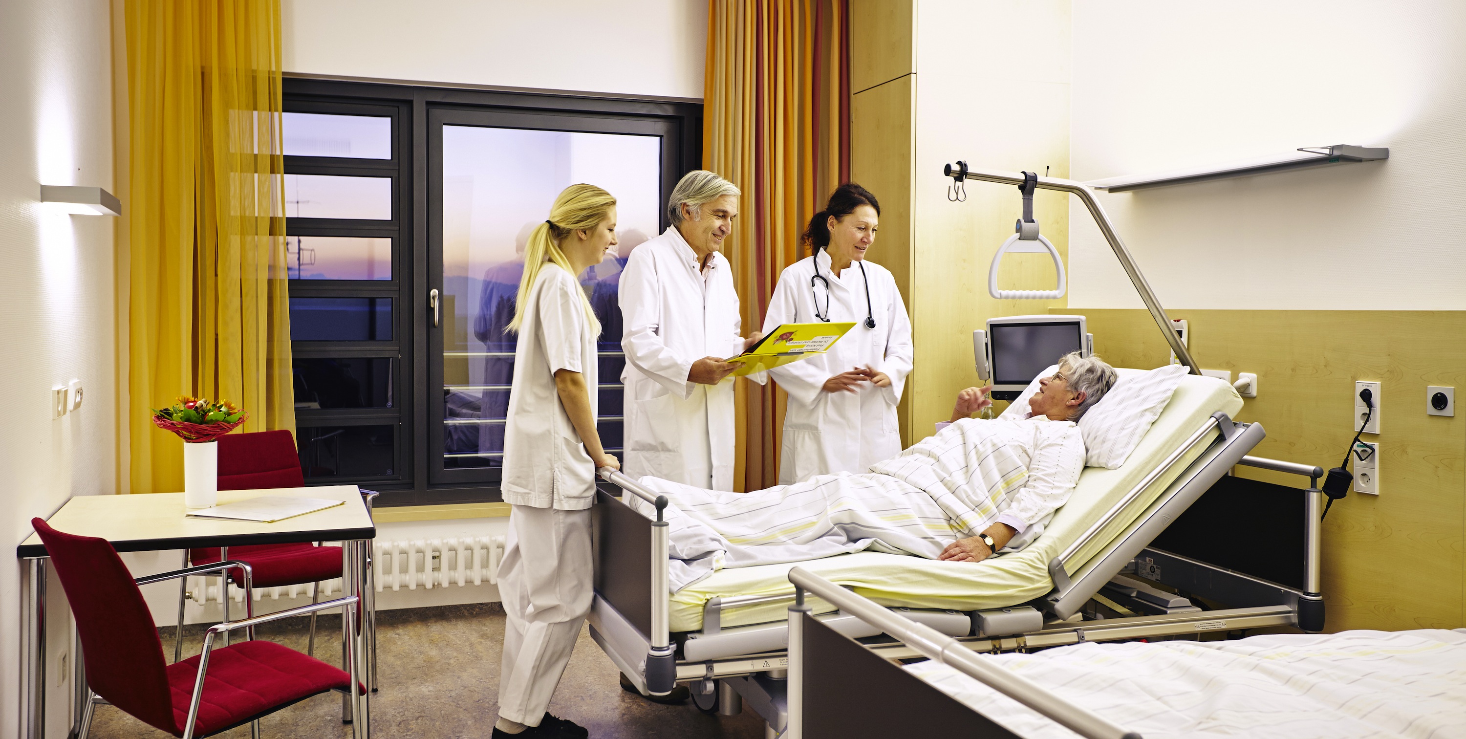3 Personen in weißen Mänteln vor einem Krankenbett im Krankenzimmer mit gelben Vorhängen 