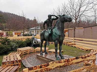 Eine oxidierte Bronzestatue eines nackten Reiters, welcher auf einem Pferd Ausschau hält und dabei sein Gesicht mit der linken Hand von der Sonne zu schützen versucht. Umringt ist er dabei von Holzpaletten.