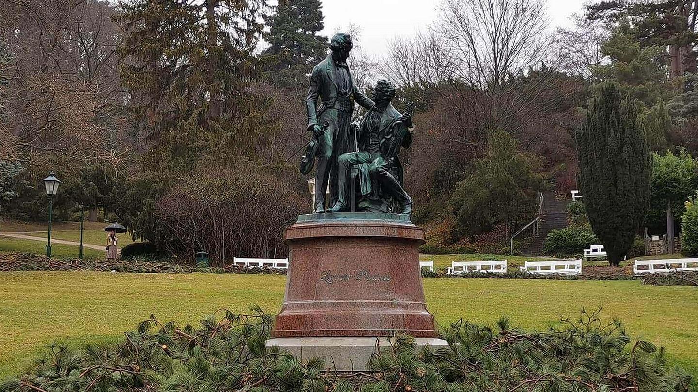 Zwei oxidierte Bronzestatuen von Joseph Lanner und Johann Strauß auf einem runden Fundament umringt von Ästen von Nadelbäumen, während im Hintergrund eine weite Wiese und Bäume zu sehen sind.