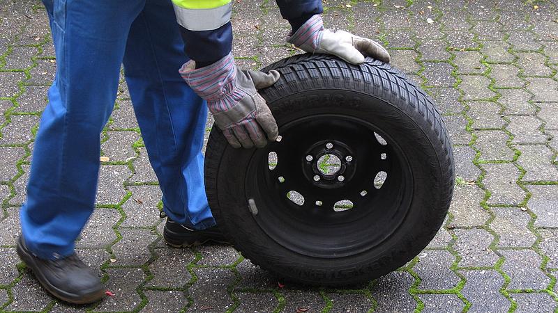 Mechaniker in Arbeitskleidung hält einen Winterreifen ins Bild.