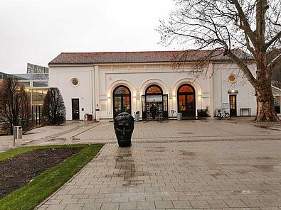 Ein großer asphaltierter Platz vor der Römertherme in Baden, während die Statue eines oxidiertetn Kaiserkopfes aus Bronze vor dem Gebäude steht.