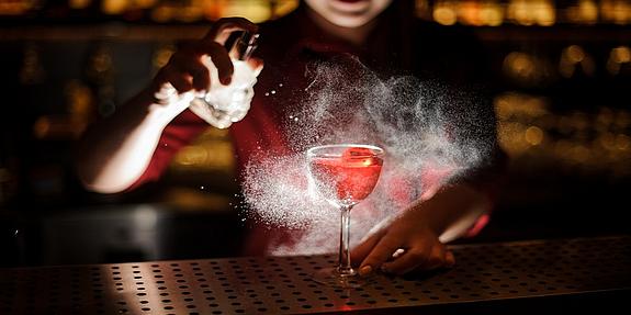 Die Barkeeperin einer Speakeasy-Bar besprüht einen Cocktail mit aromatischem Duft. Das gedämpfte Licht sorgt für eine geheimnisvolle Atmosphäre.