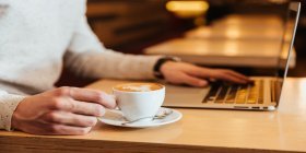 Person sitzt mit Cappucchino im Kaffeehaus und arbeitet am Laptop.