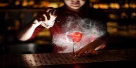 Die Barkeeperin einer Speakeasy-Bar besprüht einen Cocktail mit aromatischem Duft. Das gedämpfte Licht sorgt für eine geheimnisvolle Atmosphäre.