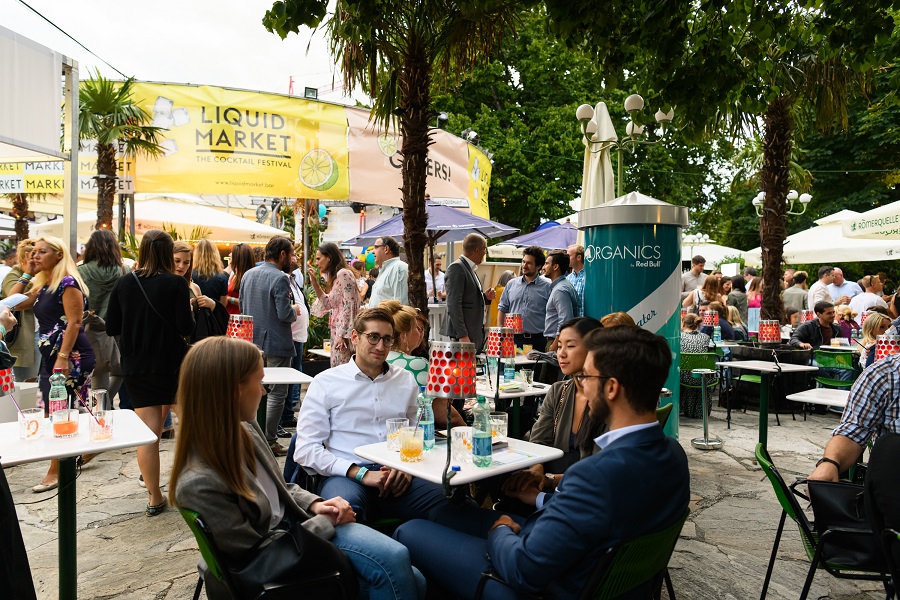 Liquid Market Logo im Hintergrund, Menschen sitzen mit Getränken an einem Tisch