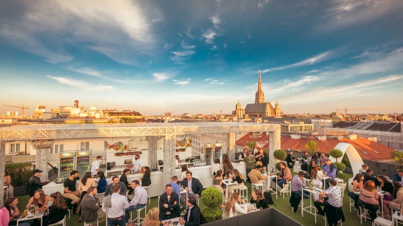 Menschen die unter blauem Himmel auf einer Dachterrasse über den Dächern Wiens sitzen.