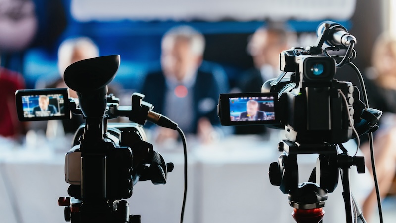 Zwei Fernsehkameras, im Hintergrund mehrere Personen bei einer Konferenz. 